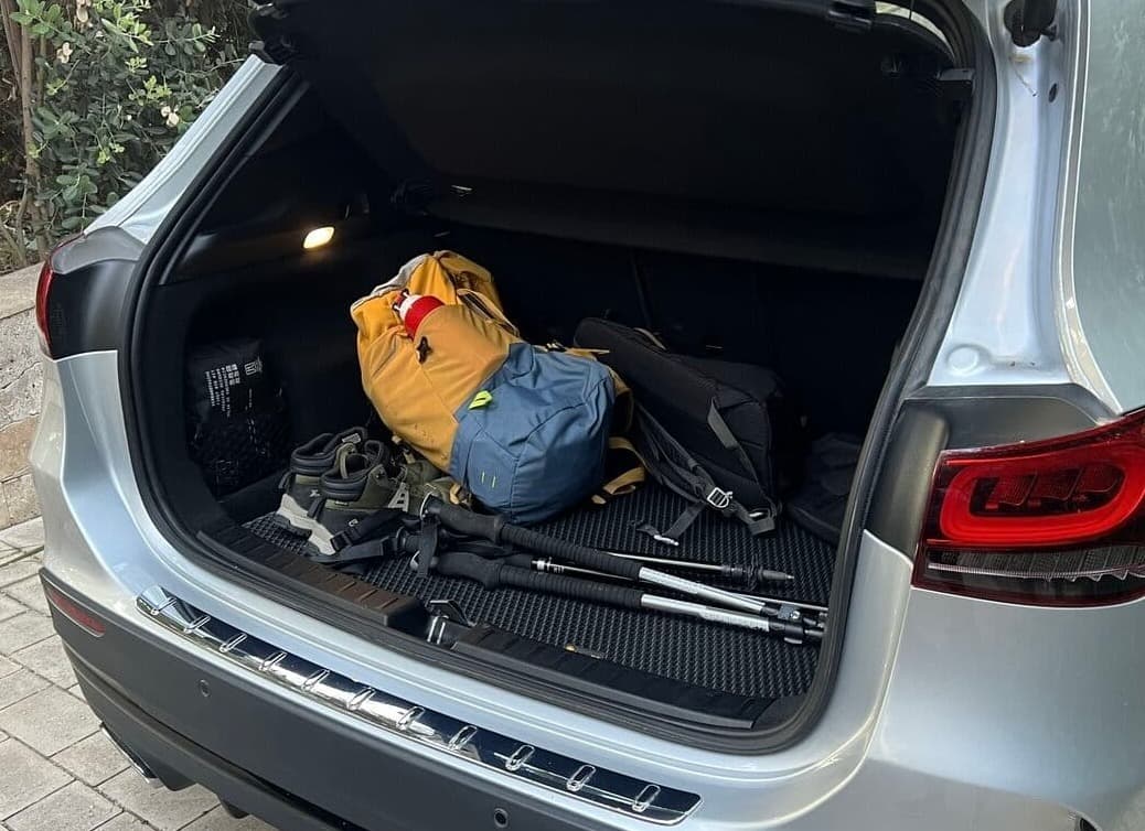 Offener Autokofferraum mit Wanderausrüstung, darunter Rucksäcke, Trekkingstöcke und Schuhe auf einer strukturierten Auto-Matte.