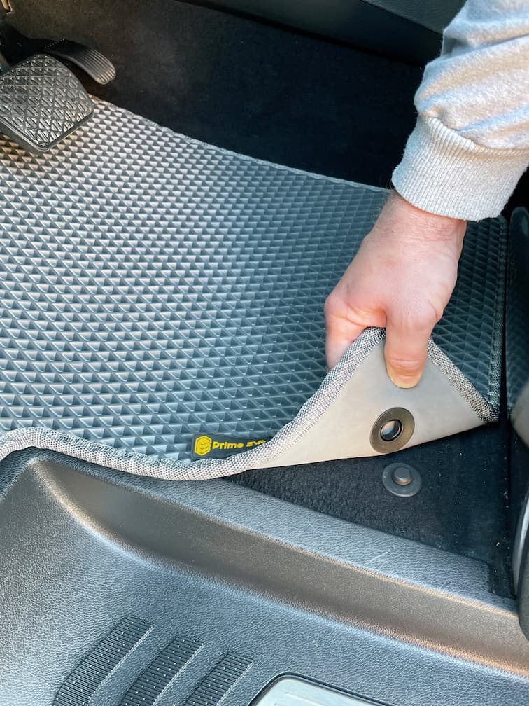 Hand hebt graue EVA-Autofußmatte an und zeigt Befestigungsdetail nahe den Pedalen.