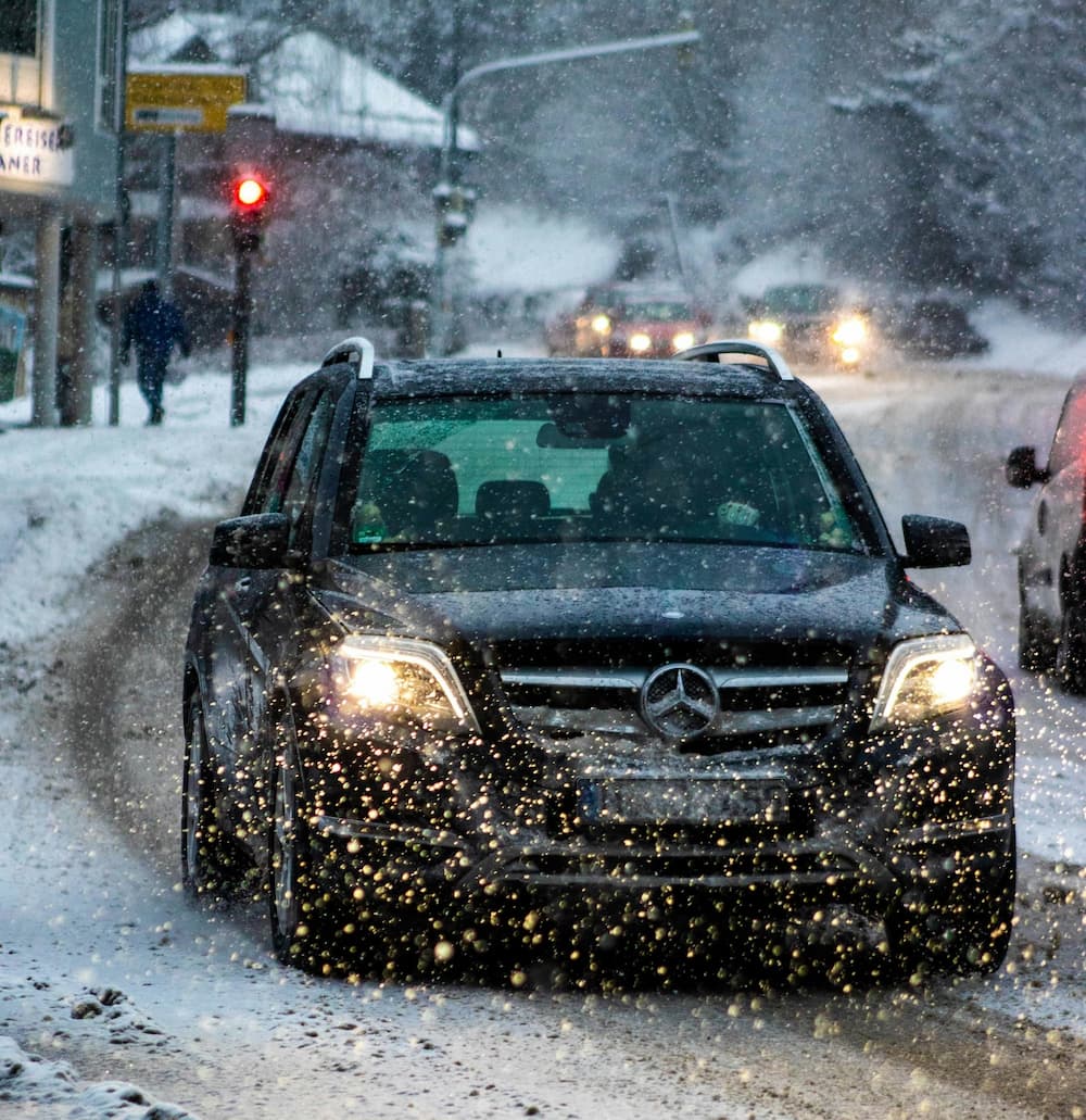 Mercedes SUV fährt mit eingeschalteten Scheinwerfern durch starken Schneefall auf winterlicher Straße.