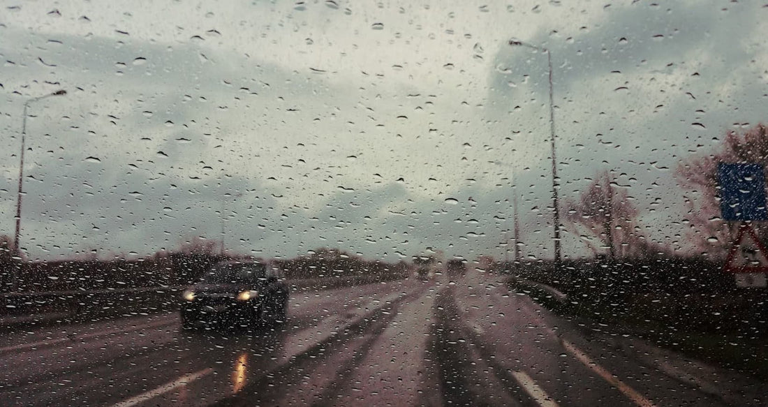 Eine regnerische Straßenansicht durch eine Windschutzscheibe mit Wassertropfen und einem unscharfen Fahrzeug voraus.