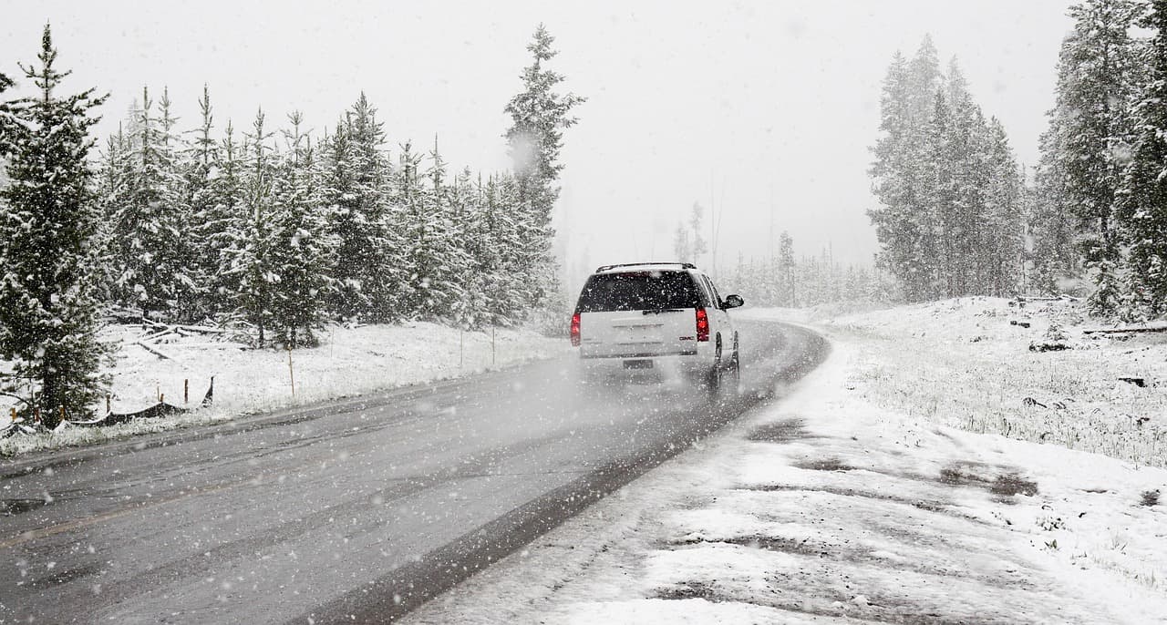 Ein weißer SUV fährt auf einer verschneiten Straße, umgeben von Kiefern, während es schneit.