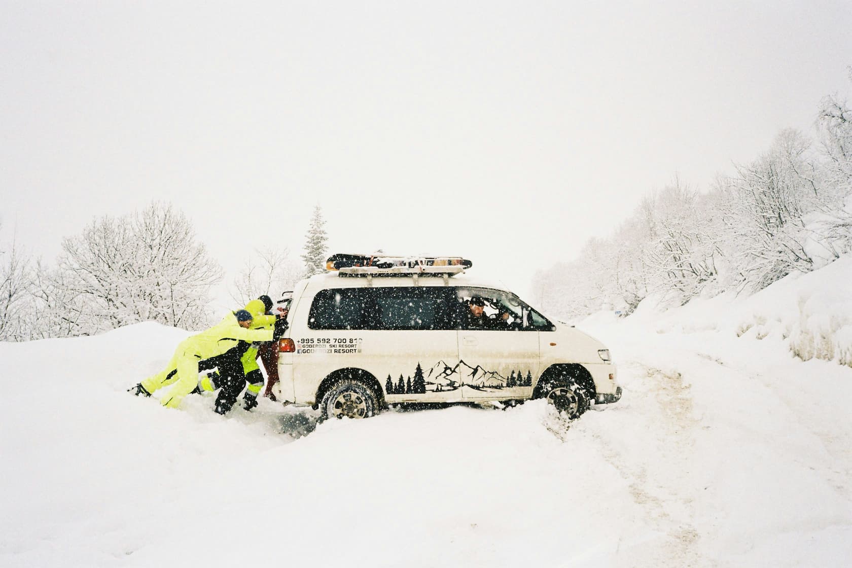 Mehrere Personen schieben einen im tiefen Schnee festgefahrenen Van auf winterlicher Bergstrasse.